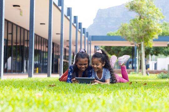 Two biracial girls are engrossed in a tablet outdoors at school, lying on the grass