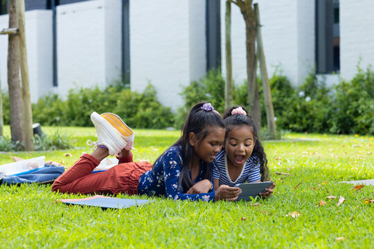Two biracial girls are lying on the grass in school, engaged with a smartphone
