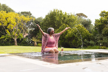 Senior biracial woman enjoys a playful moment by a pool, splashing water with her hands, with copy s