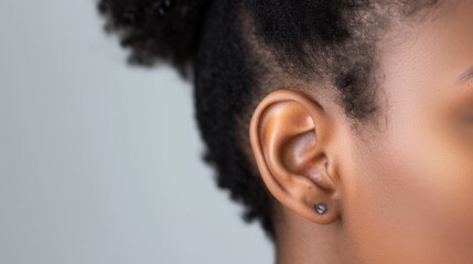 The image shows a close-up of a young womans ear. The woman has dark skin and her hair is styled in a bun. She is wearing a silver earring in her left ear. The background is a soft, light gray. - obrazy, fototapety, plakaty