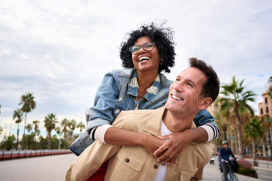 Mature multiracial joyful tourist couple piggybacking standing outdoor. Cheerful black woman and Caucasian man smiling walking on city street enjoying happy vacation. Copy space, background palm trees