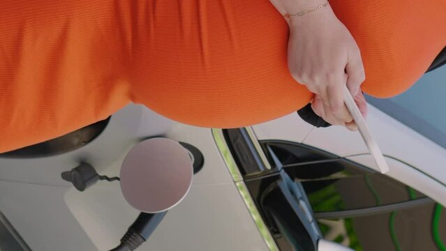 Vertical video of unrecognizable pregnant woman in orange dress using the internet on her smartphone while waiting charging the battery at the electric car