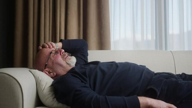 Elderly man with glasses lying on the sofa at home has a headache and rubs his head.