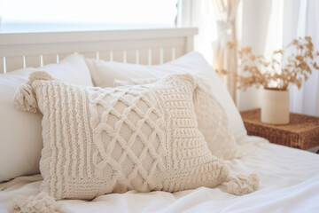 Closeup of the white knitted pillow with brushes on a bed with a white wooden headboard, dried flower in a vase on the bedside table.