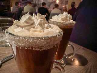 Clear glass mugs of Irish coffee with whipped cream and sugared rims. The tall glass is on a wooden bar at a pub. The white thick cream is on top of the hot coffee and strong whisky.