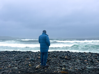 A man standing on a beach wearing a blue winter coat and hat looking towards a stormy ocean. The beach is covered in large rocks. The cold sea is rough with waves crashing on the shoreline.
