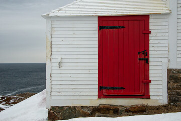 The corner of a white wooden shed covered with narrow clapboard siding. There's a wide red door with black hinges and metal latches. The calm blue ocean is in the background with snow on the ground.