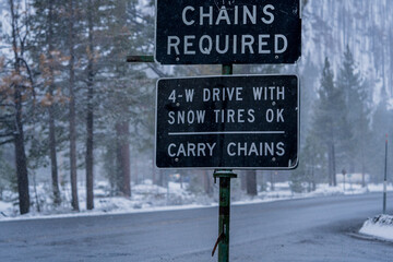 Highway sign in Sierra Nevada mountains that reads,"Chains Required -- 4-W Drive with Snow Tires OK -- Carry Chains"  Snowy winter backdrop with wet road.  Captured during a winter storm.  