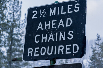 Highway sign in Sierra Nevada mountains that reads,"Two and a half Miles Ahead Chains Required"  Snowy winter backdrop with trees.  Captured during the onset of a winter storm.  