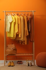 Rack with different stylish women's clothes, shoes, backpack and pouf near orange wall indoors