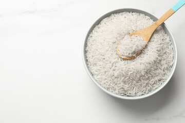 Raw basmati rice with spoon in bowl on white marble table, top view. Space for text