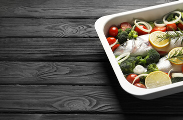 Raw fish with vegetables and lemon in baking dish on black wooden table. Space for text