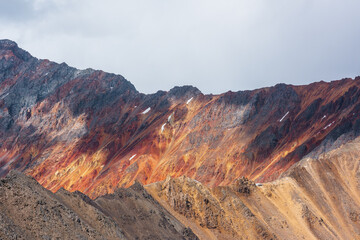 Colorful layered landscape with sunlit sharp rocky mountain ridge of vivid colors under cloudy sky. Golden rocks and red sheer crags in sunlight. Shadows of clouds on unusual multicolored mountains.