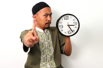 Indonesian Muslim man in koko and peci holds a clock and raises his finger, signaling just a little...