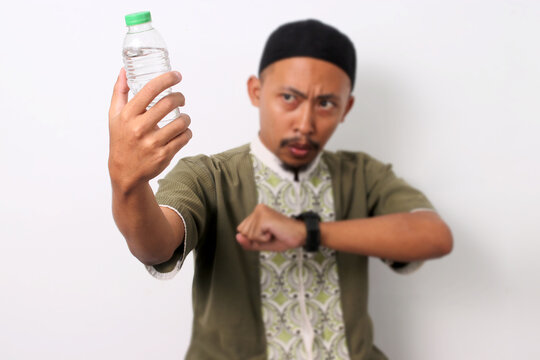 An Indonesian Muslim man in koko and peci checks his watch, waiting for iftar during the Ramadan fast. He holds a water bottle, indicating his thirst. Isolated on a white background