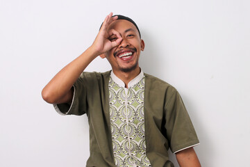 Happy Indonesian Muslim man in koko and peci playfully makes an OK gesture near his eyes, mimicking...