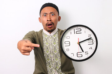 A shocked Indonesian Muslim man in koko and peci emphatically points at a clock, realizing he's late for his sahur meal during Ramadan. Isolated on a white background - obrazy, fototapety, plakaty