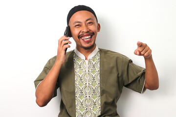 Indonesian Muslim man in koko and peci points his finger emphatically at camera while talking on his phone during Ramadan. Isolated on a white background - obrazy, fototapety, plakaty