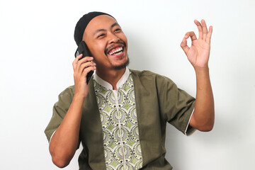 Happy Indonesian Muslim man in koko and peci makes an OK gesture while talking on his phone,...
