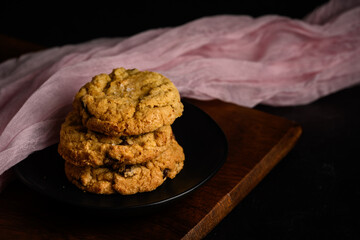 Spring treat, dark and moody image of three homemade fancy chocolate chip cookies stacked on a black plate with pale pink gauze clothe behind
