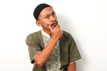 A pensive Indonesian Muslim man in koko and peci rests his hand on his chin, gazing thoughtfully at the camera during Ramadan. Isolated on a white background