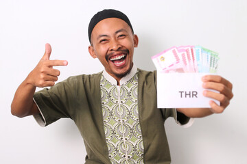 Excited Indonesian Muslim man points his finger at a white envelope labeled THR, filled with...