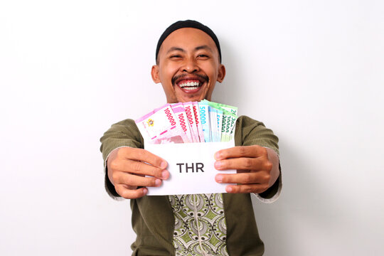 An excited Indonesian Muslim man in koko and peci holds a white envelope labeled THR, filled with money, representing his Religious Holiday Allowance. Isolated on a white background