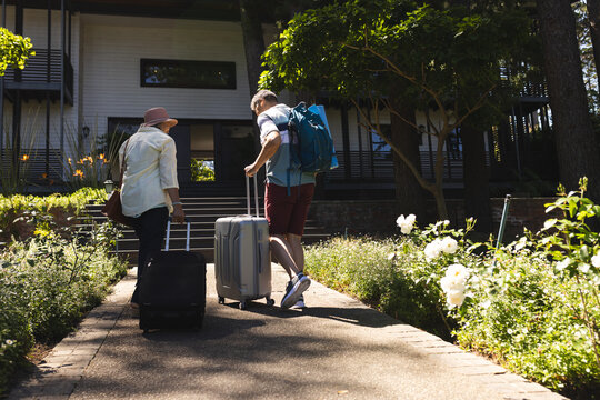 Biracial couple walks towards a house, pulling a suitcase and carrying a backpack, with copy space