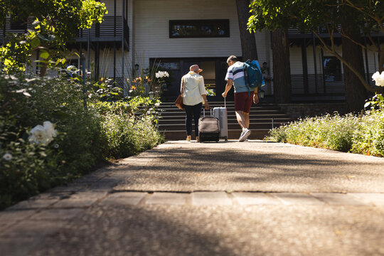 Senior biracial woman and biracial man walk towards a house, pulling a suitcase, with copy space