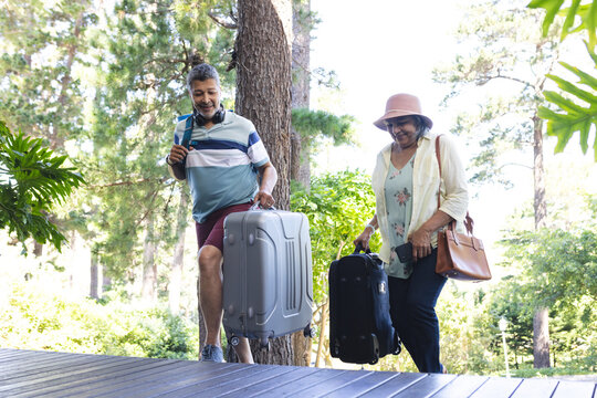 Senior biracial couple walking with luggage in a lush park setting