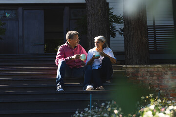 Senior biracial woman and biracial man sit on steps, enjoying a conversation with coffee