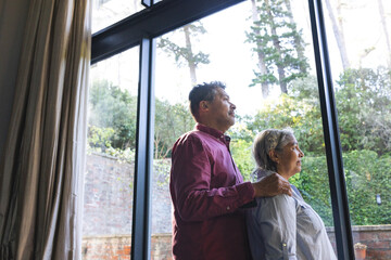 Senior biracial couple stands by a large window, looking outside thoughtfully