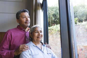 Senior biracial couple stands by a window with copy space, the woman smiling gently, the man looking