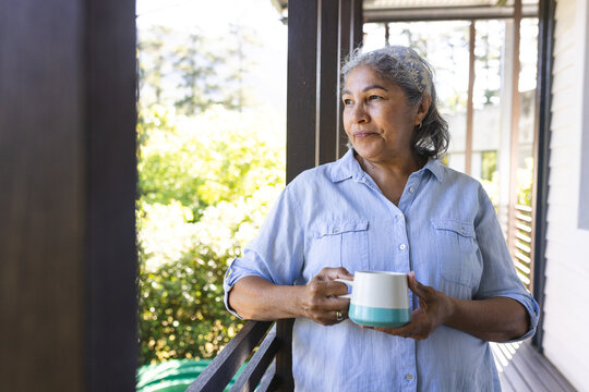 Senior biracial woman with grey hair stands on a porch, holding a mug, with copy space - Powered by Adobe