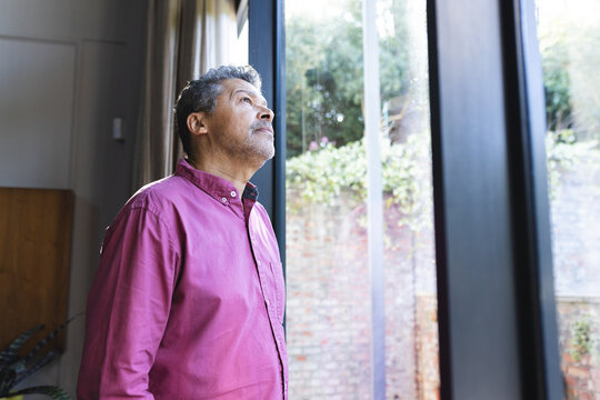 Senior biracial man in a pink shirt looks contemplatively out a window with copy space