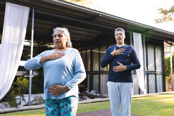 Senior biracial woman and biracial man practice yoga outdoors, both with hands on their chests, at h