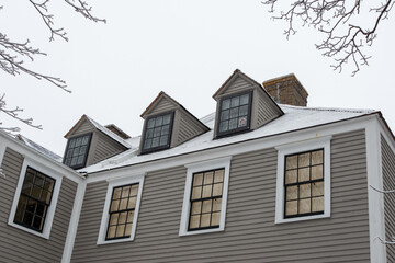 A vintage tan color wooden building with white trim, multiple double-hung windows, dormers, gables, a black shingled roof, and a brown brick chimney. The peaked roof is covered in fresh white snow.