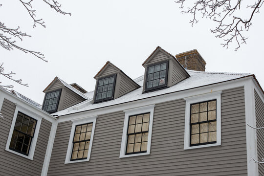 A vintage tan color wooden building with white trim, multiple double-hung windows, dormers, gables, a black shingled roof, and a brown brick chimney. The peaked roof is covered in fresh white snow.
