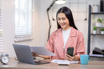 Portrait of happy young asian woman accountant working documents on laptop and  phone desk using calculator for calculate finance report in home office