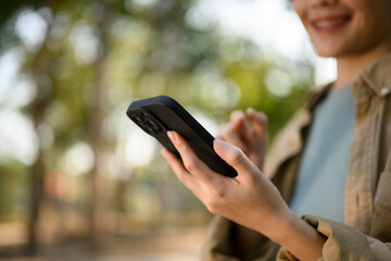 Smiling young woman typing message on mobile phone, standing in the public park