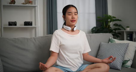 Happy young woman wear automatic massage neck meditating doing a lotus pose at home with eyes...