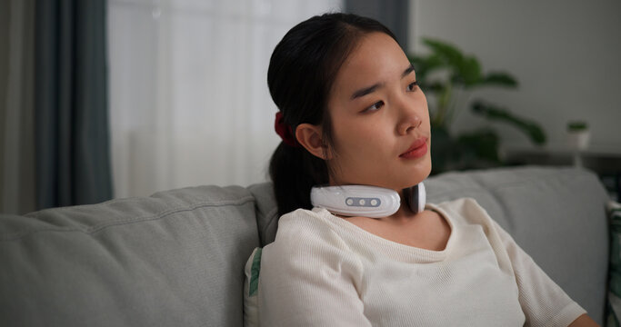 Selective focus, Relaxed young woman wear automatic massage neck sitting on sofa at home ,relaxing body and mind alone in the living room, breathing fresh air,