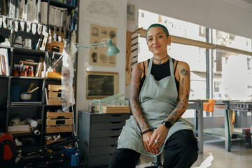 Smiling female designer sitting on background of own workshop and looks camera