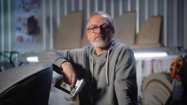 Professional carpenter stands, holds upholstery stapler and looks at camera. Mature craftsman works in workshop for assembling, making or restoring furniture. Handcraft and small business. Portrait.