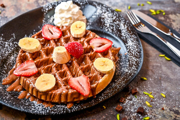 Strawberry and Banana waffle with chocolate, whipped cream, pistachio, knife and fork served in dish isolated on dark background top view cafe dessert food