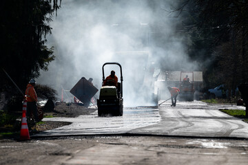 Residential street repaving project, silhouetted steamroller compacting and smoothing new asphalt,...