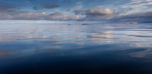 Calm blue sea, blue sky and clouds; summer haze on the surface of the water. some rocks, islets, islands and mountains in the background. Lofoten Islands, Northern Norway.