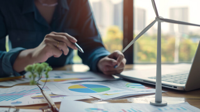 person analyzing sustainability data, with a small model wind turbine in the foreground, symbolizing renewable energy. - Powered by Adobe
