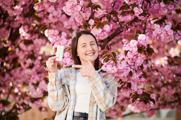Fototapeta premium Woman allergic suffering from seasonal allergy at spring. Young happy woman showing pack of pills, posing in blossoming garden at springtime. Antihistamine medication concept