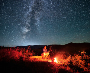 Man taking photos of starry sky with Milky way. Male photographer sitting on log near camera. Man...